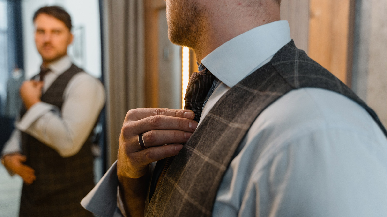 A man in a suit and waistcoat is adjusting his tie in front of a mirror, with a tungsten ring on his hand